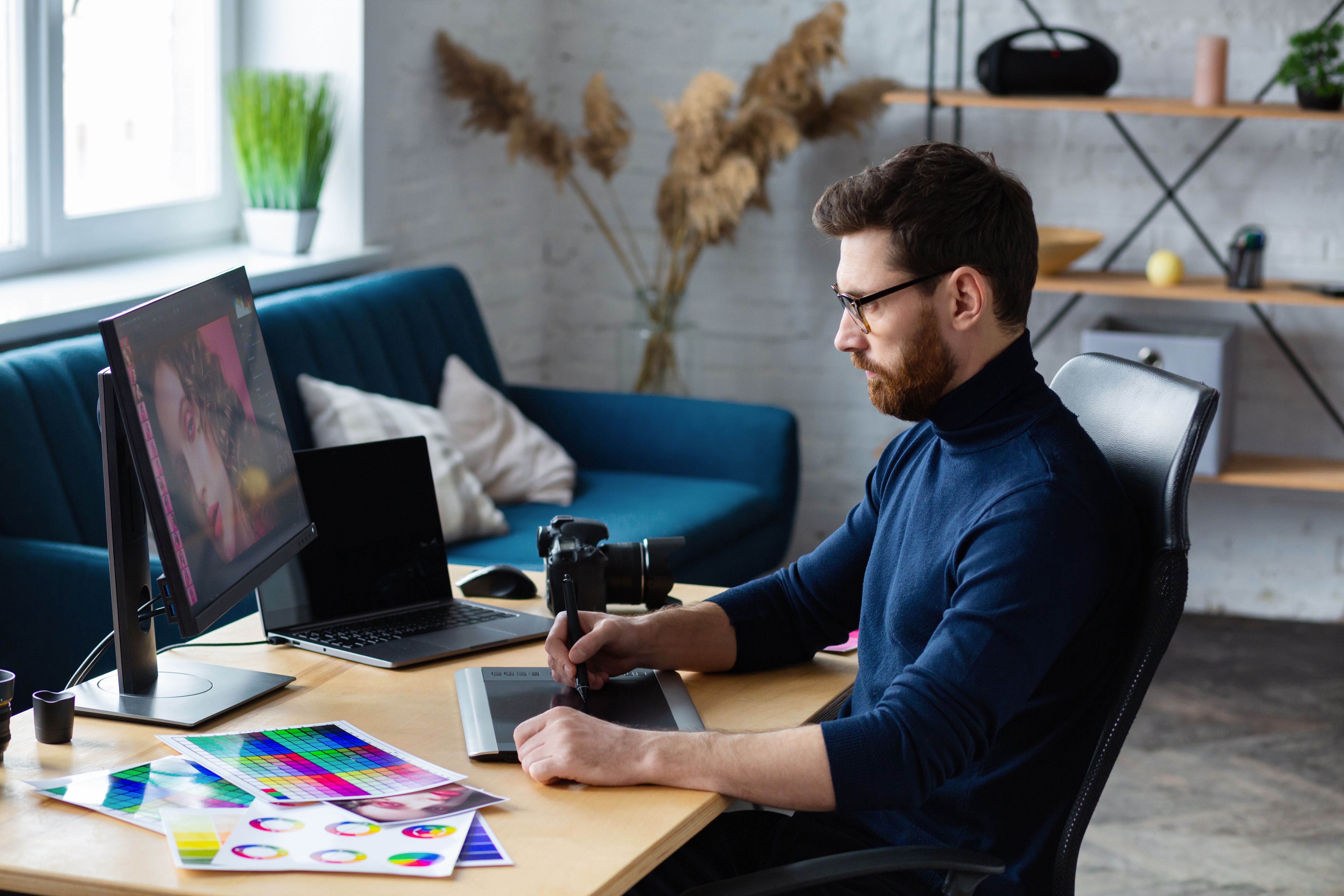 Profesjonalny graphic designer pracujący przy biurku z laptopem, monitorem z portretem, tabletem graficznym Wacom, aparatem DSLR i próbnikiem kolorów Pantone