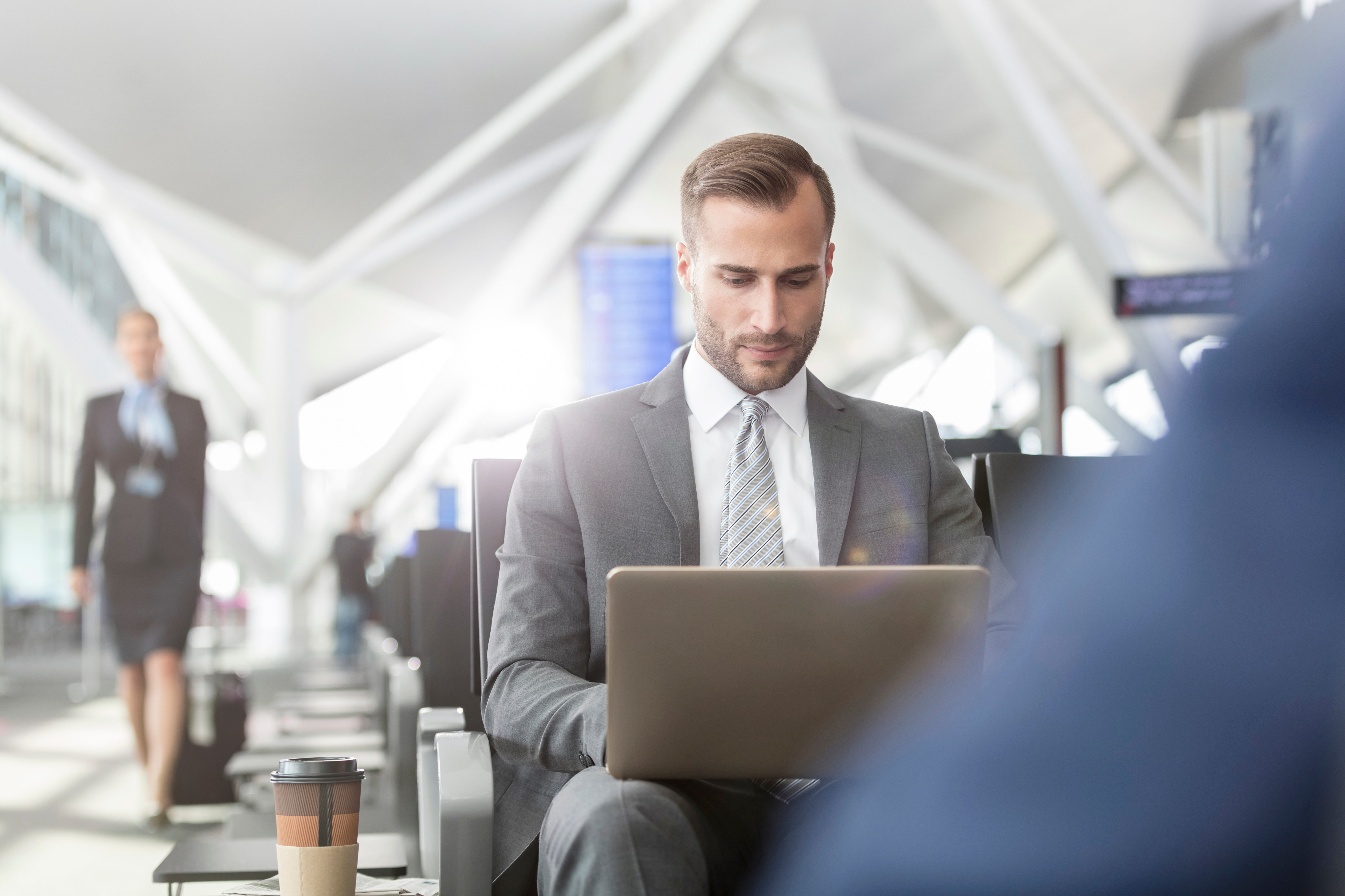 Businessman w garniturze pracujący na laptopie w hali odlotów lotniska, z kawą obok, w tle inni pasażerowie